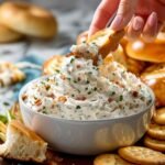 Hand dipping a cracker into a bowl of creamy bagel dip garnished with bacon and herbs.