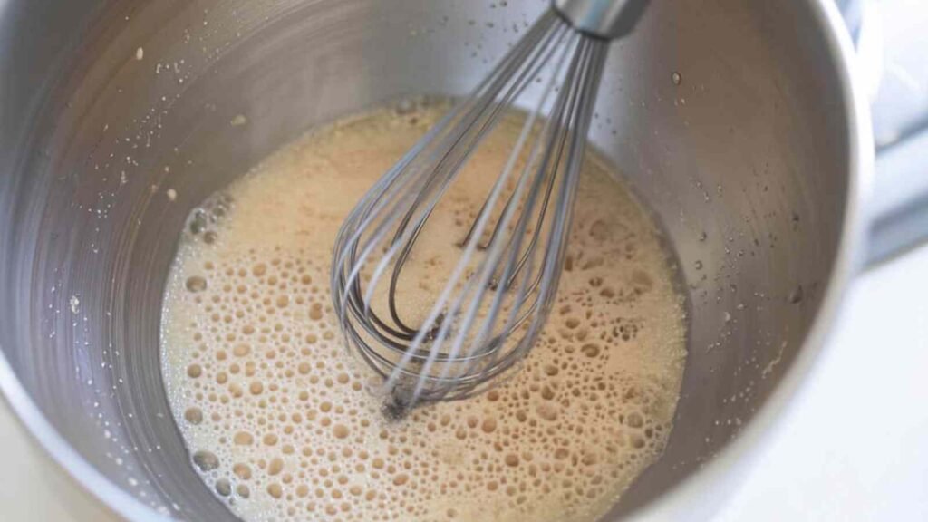 Whisk stirring foamy yeast mixture in a metal bowl as the first step to making sourdough discard bagels.
