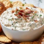 Bagel dip garnished with bacon and herbs on a wooden platter, surrounded by pretzels and crackers.