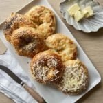 Platter of assorted golden cottage cheese bagels with shredded cheese and poppy seeds, served with butter on the side.