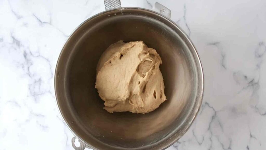 Ball of kneaded bagel dough in a metal mixing bowl, ready for overnight rise on a marble countertop.