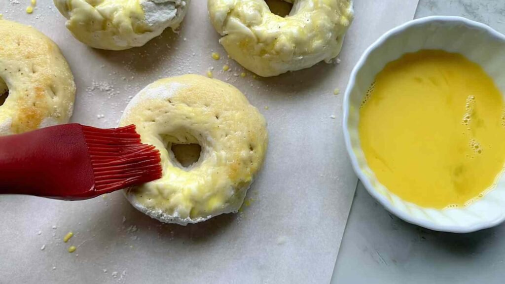 Silicone brush applying egg wash to raw bagel dough on parchment paper next to a bowl of whisked egg.