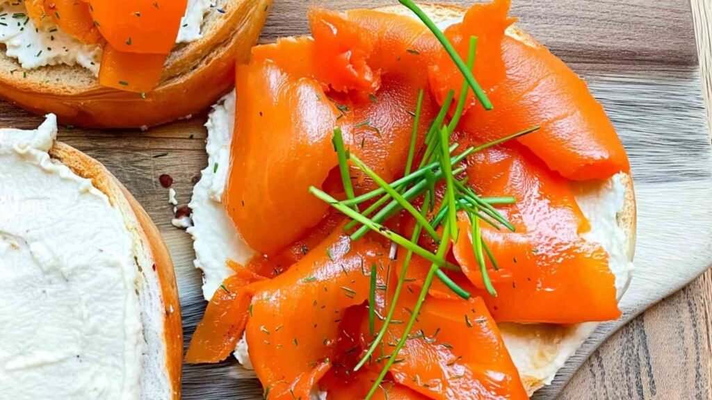 Close-up of a toasted bagel topped with creamy vegan cheese, smoky carrot lox ribbons, and fresh chives on a wooden board.