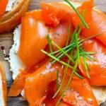 Close-up of a toasted bagel topped with creamy vegan cheese, smoky carrot lox ribbons, and fresh chives on a wooden board.