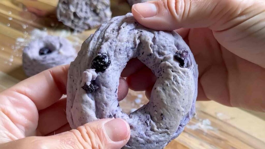 Hands shaping a raw blueberry bagel dough ring, with visible berries and a soft texture.