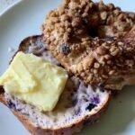 Greek yogurt blueberry bagel sliced open on a plate, topped with a pat of melting butter and a golden cinnamon crumble.