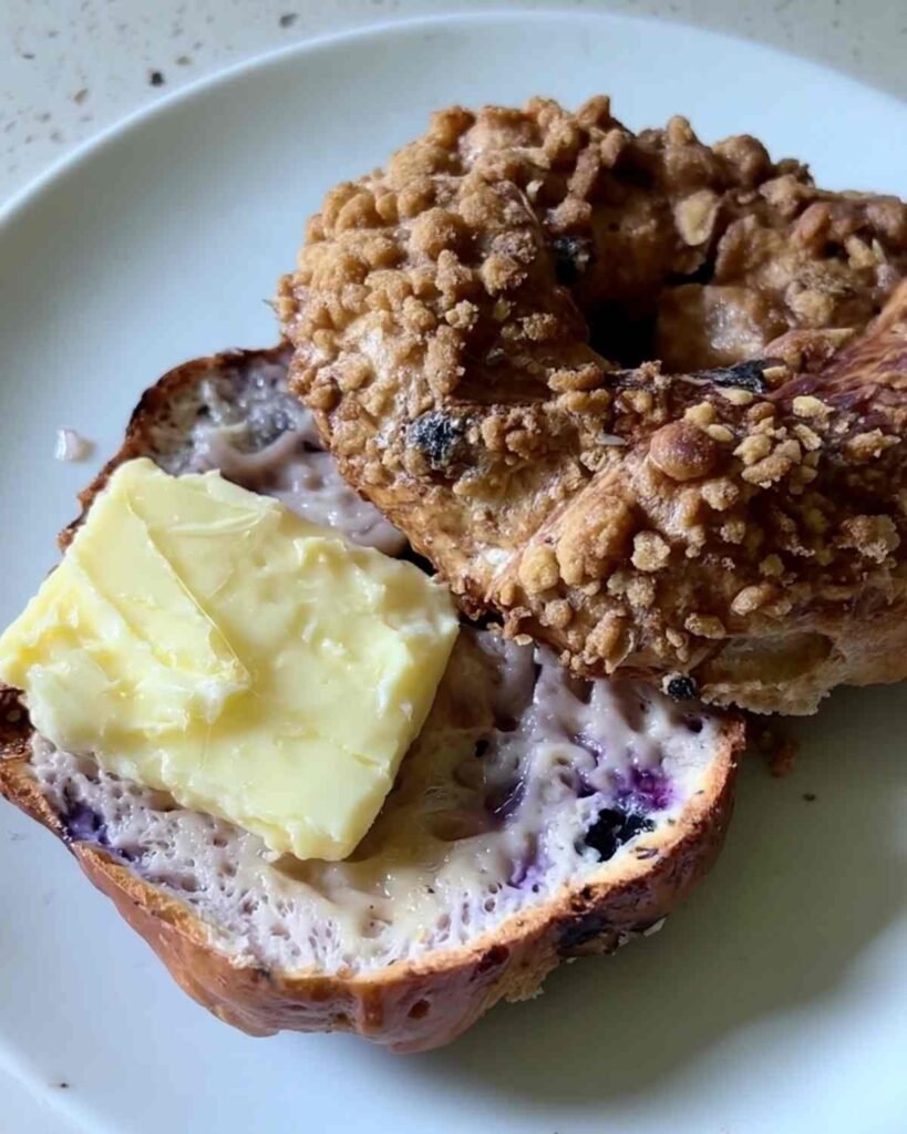 Greek yogurt blueberry bagel sliced open on a plate, topped with a pat of melting butter and a golden cinnamon crumble.