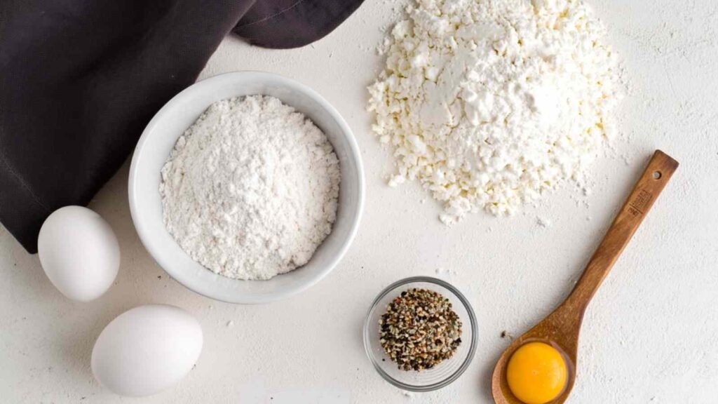 Top-down view of self-rising flour, full-fat cottage cheese, eggs, seasoning blend, and egg yolk on a white surface for making 3 ingredient cottage cheese bagels

