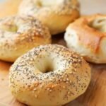 Golden brown baked keto high-protein bagels on a wooden board, topped with sesame seeds and poppy seeds, showing chewy crust and soft center