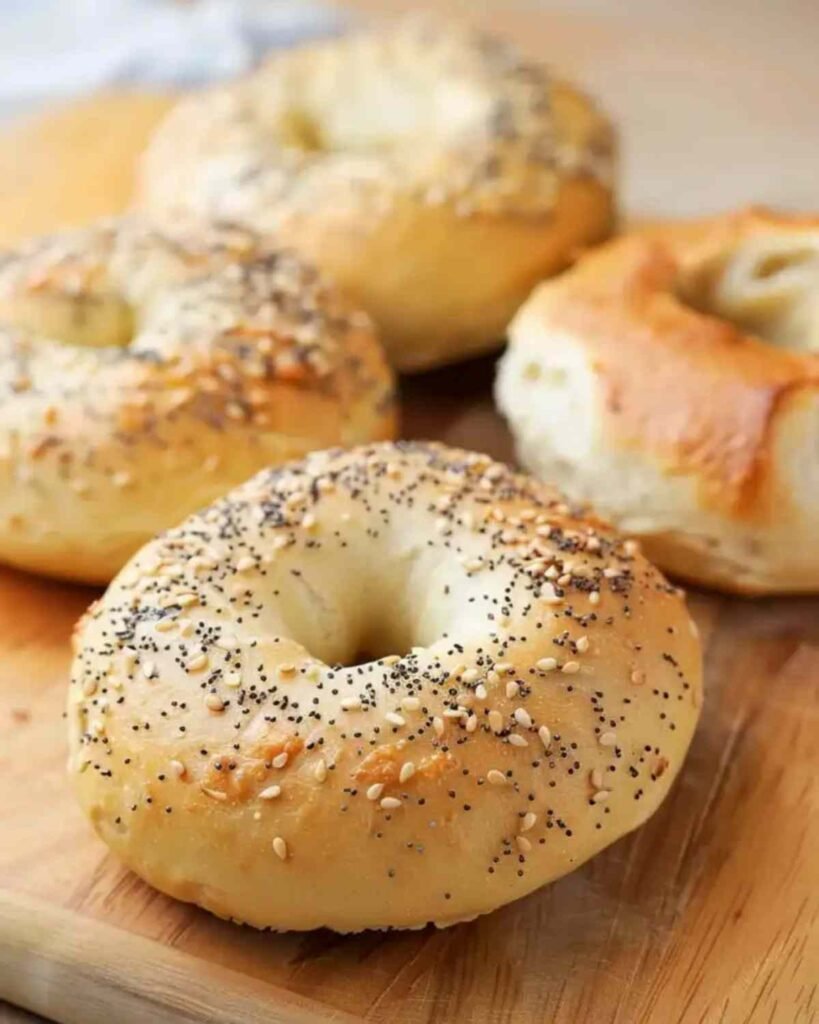 Golden brown baked keto high-protein bagels on a wooden board, topped with sesame seeds and poppy seeds, showing chewy crust and soft center

