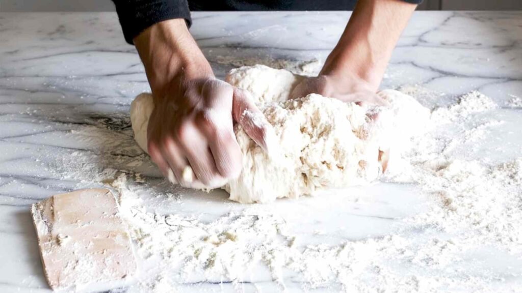 Hands kneading stiff bagel dough on a floured marble countertop, prepping for chewy New York Style Bagels