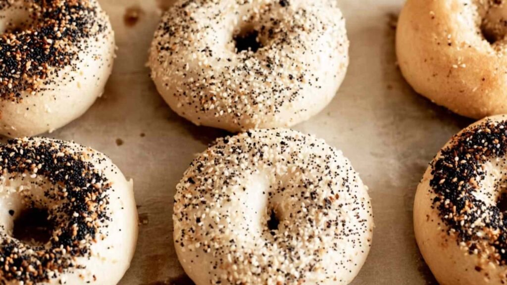 Unbaked everything bagels resting on a baking sheet, coated with poppy, sesame, and garlic flakes
