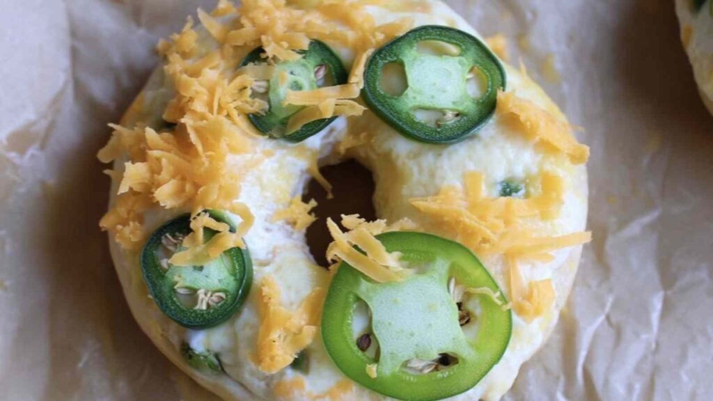 Close-up of raw jalapeno cheddar bagel topped with fresh sliced jalapeños and shredded cheddar cheese on parchment paper.