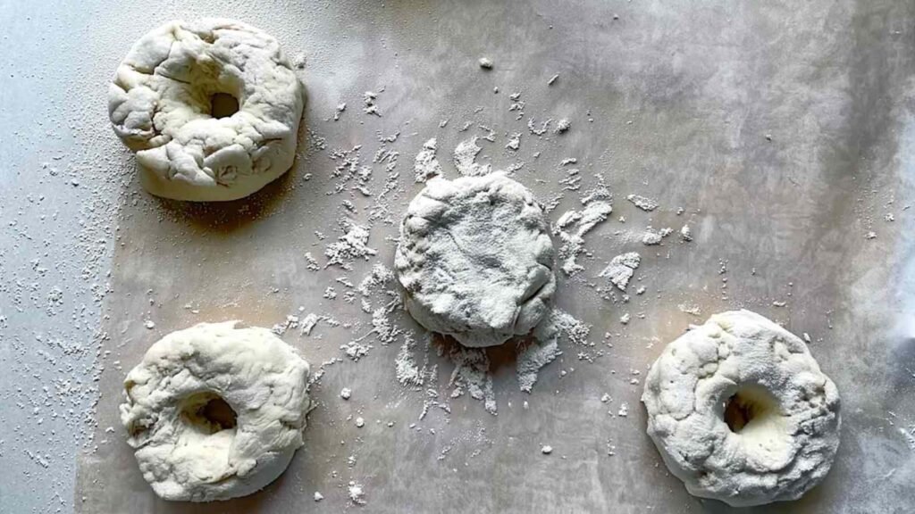 Floured parchment paper with four shaped but unbaked cottage cheese bagels and scattered flour.