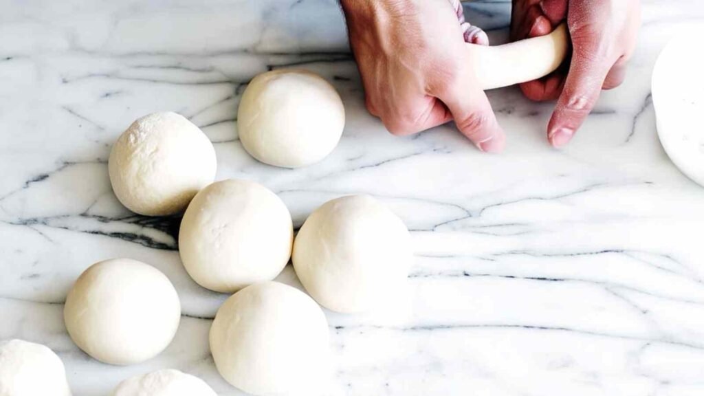 Hands stretching a ball of bagel dough into a ring, with smooth dough balls resting on a marble surface