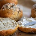 Sliced sourdough discard bagel with cream cheese being spread on one half, surrounded by whole bagels.