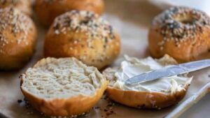 Sliced sourdough discard bagel with cream cheese being spread on one half, surrounded by whole bagels.