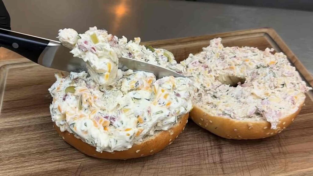 Knife spreading a thick, creamy mixture of chopped pickles, cheddar, onion, and herbs onto a toasted sesame bagel.
