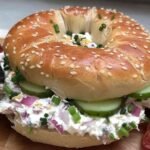 Sesame bagel filled with colorful layers of chopped pickle mix, cucumbers, red onions, shredded cheddar, and bacon strips.