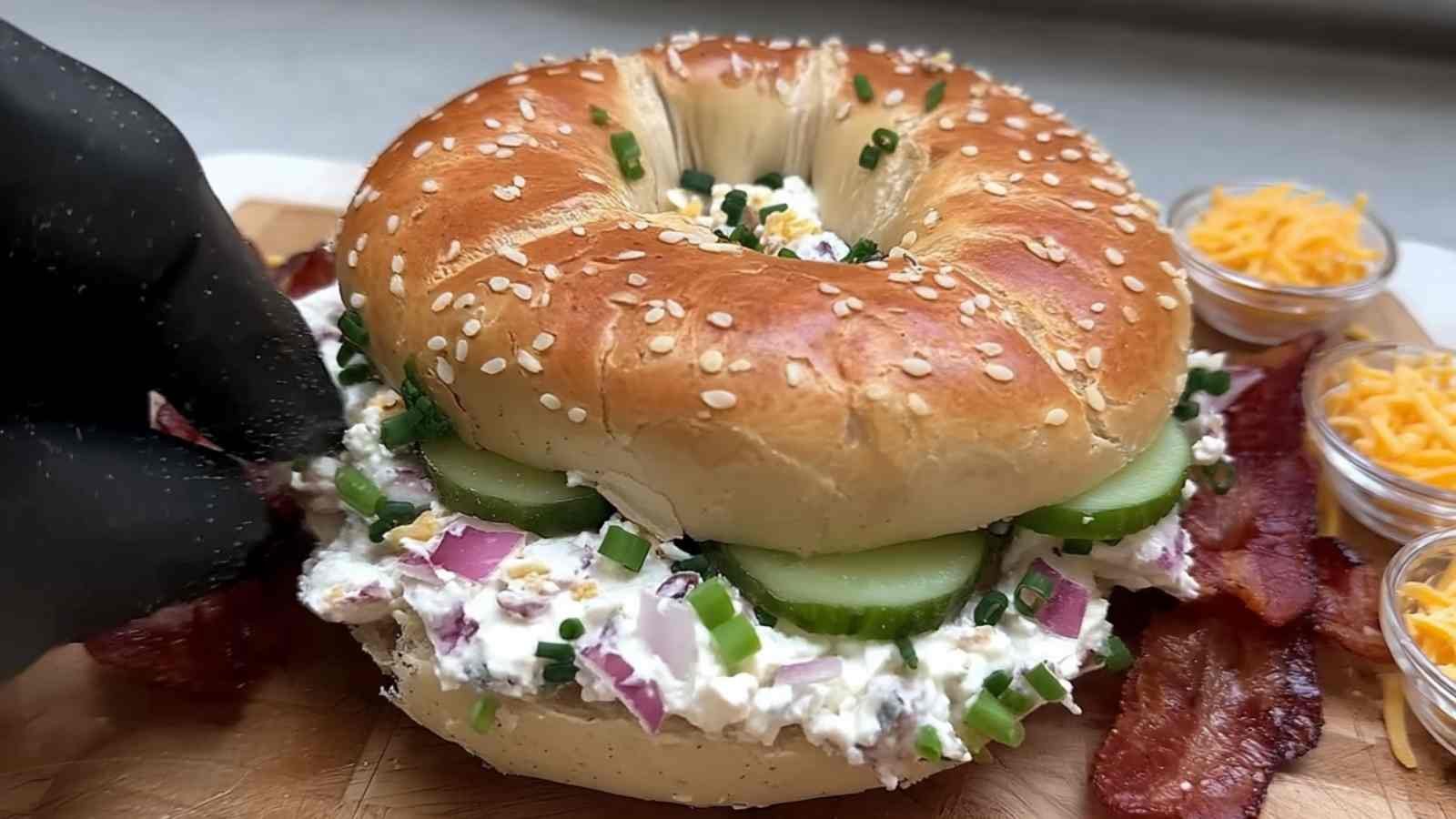 Sesame bagel filled with colorful layers of chopped pickle mix, cucumbers, red onions, shredded cheddar, and bacon strips.