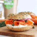 Side view of a sesame bagel stacked with vegan cream cheese, carrot lox, red onions, and capers on a cutting board.