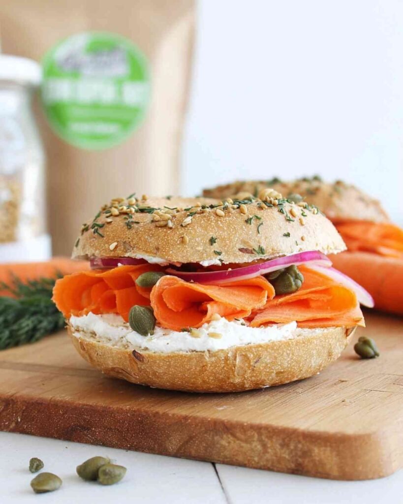 Side view of a sesame bagel stacked with vegan cream cheese, carrot lox, red onions, and capers on a cutting board.