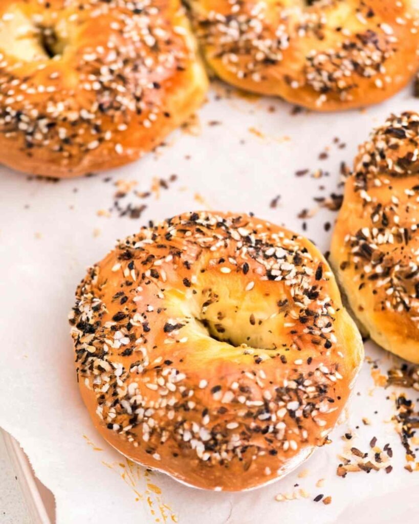 Five golden brown cottage cheese bagels topped with everything seasoning on a parchment-lined baking tray

