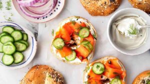 Flat lay of vegan lox bagels with sliced cucumbers, red onions, dill, and cream cheese in small bowls.