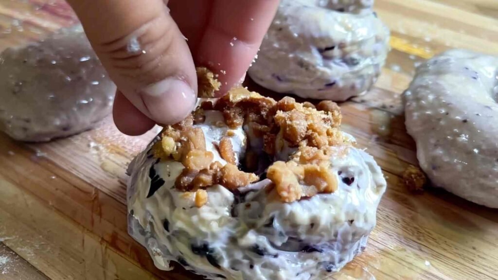 Fingers pressing cinnamon-sugar crunch topping onto unbaked blueberry bagel dough.