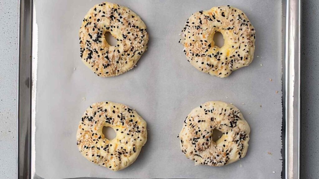 Four uncooked cottage cheese bagels on parchment paper with seasoning, placed on a baking tray.