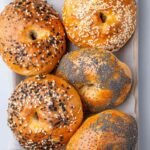 Five finished cottage cheese bagels with mixed toppings arranged in a parchment-lined tray.