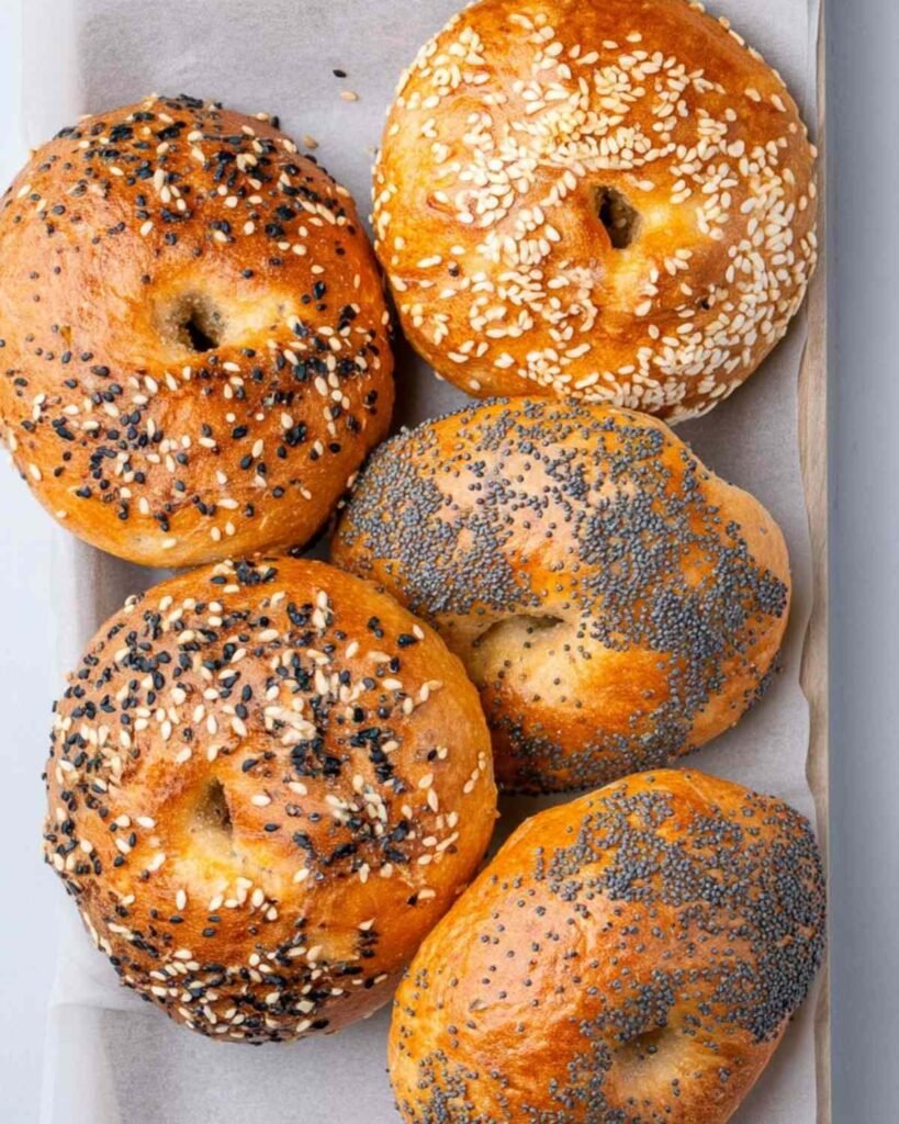 Five finished cottage cheese bagels with mixed toppings arranged in a parchment-lined tray.