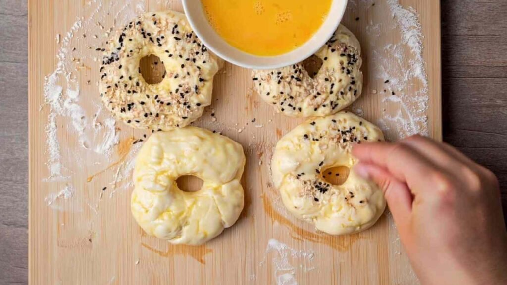 Unbaked cottage cheese bagels being topped and brushed with egg wash on a floured wooden board.