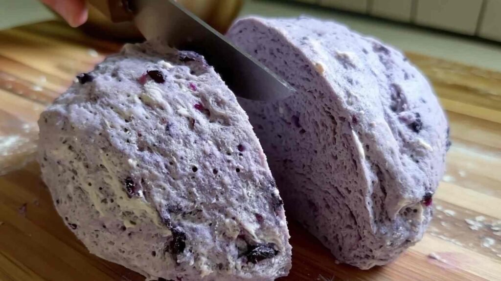 Close-up of a knife slicing a mound of blueberry-speckled dough in half on a wooden board.