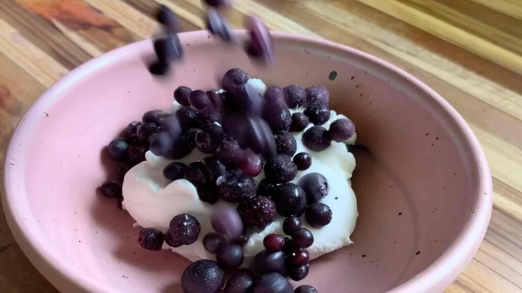 Frozen wild blueberries falling into a pink bowl of full-fat Greek yogurt before mixing into bagel dough.