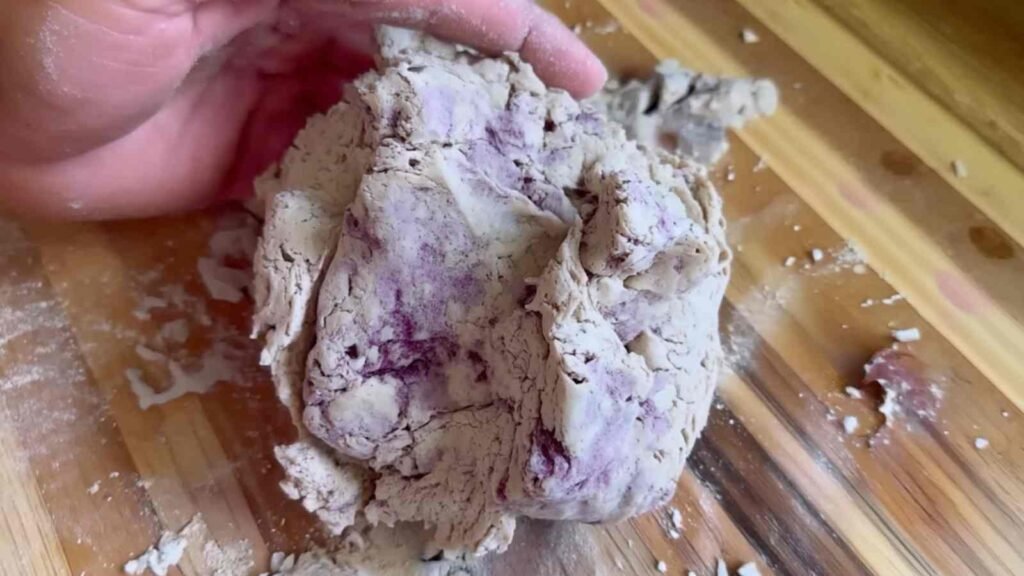 Hands kneading shaggy purple-flecked dough on a wooden surface for Greek yogurt blueberry bagels.