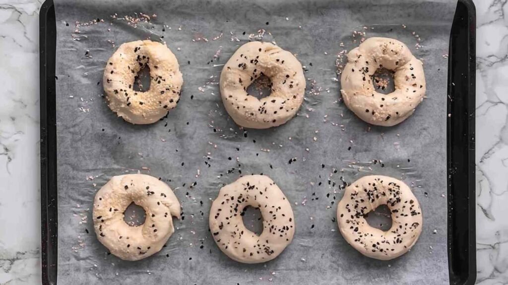 Six unbaked bagels topped with seasoning, arranged on a parchment-lined baking sheet ready for the oven.