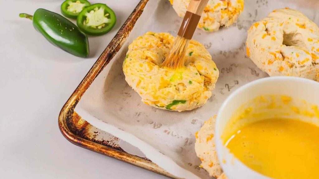 A brush applying egg wash to raw gluten-free jalapeño cheddar bagels on a baking sheet, surrounded by jalapeño slices
