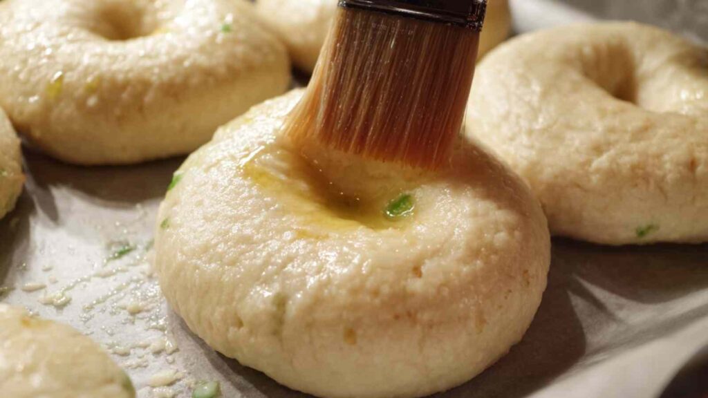 Close-up of a pastry brush applying oil to raw jalapeño bagels on a baking sheet, prepping them for the oven