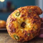 Close-up of a golden-brown gluten-free jalapeño cheddar bagel standing upright on a rustic wood table, cheese and jalapeño slices baked into the crust