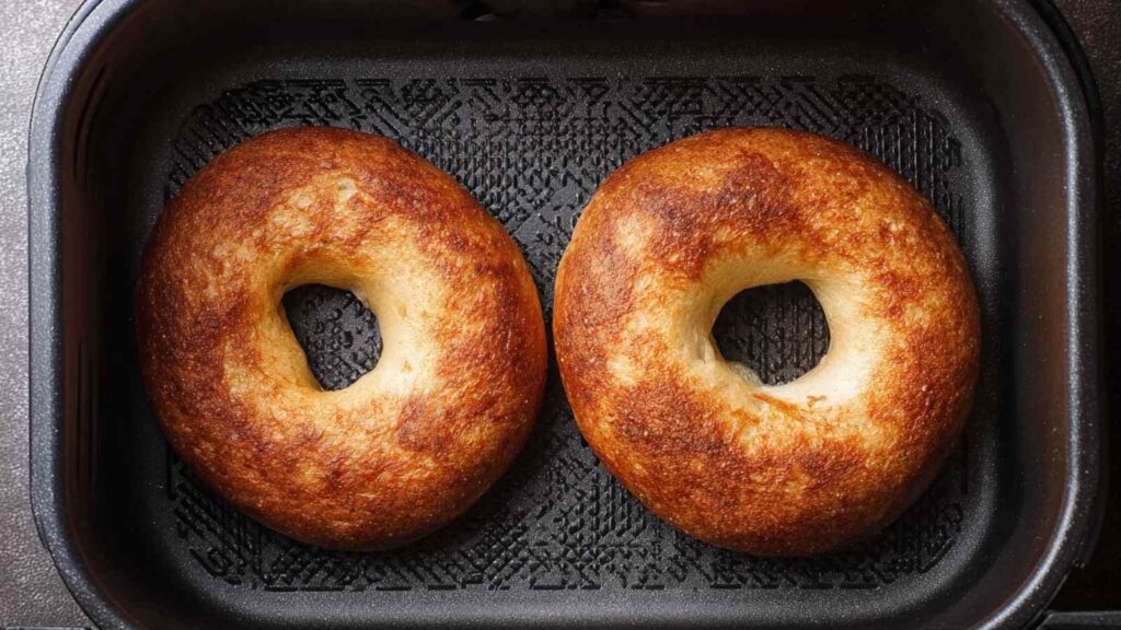 Two halved bagels toasted in the air fryer basket, golden brown and ready for toppings.