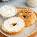 Air-fried toasted bagel halves with cream cheese on a plate beside a coffee mug and an air fryer