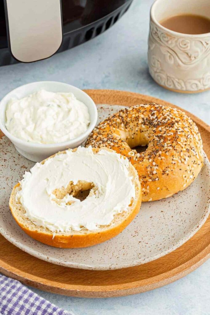 Air-fried toasted bagel halves with cream cheese on a plate beside a coffee mug and an air fryer