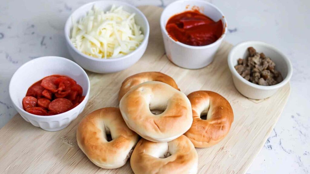 Flatlay of mini bagels, shredded mozzarella, marinara sauce, pepperoni slices, and sausage in bowls on a wooden board.