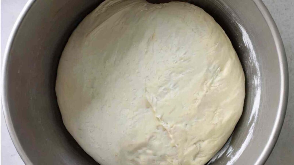 Risen dough in a metal bowl, smooth and puffed up after proofing.