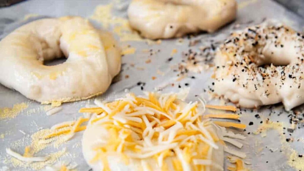 Raw shaped bagels topped with shredded cheese and seasoning, ready to be baked