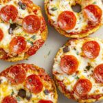 Baked mini pizza bagels on a metal tray, topped with melted cheese, pepperoni, and seasoning, with crisp golden edges.