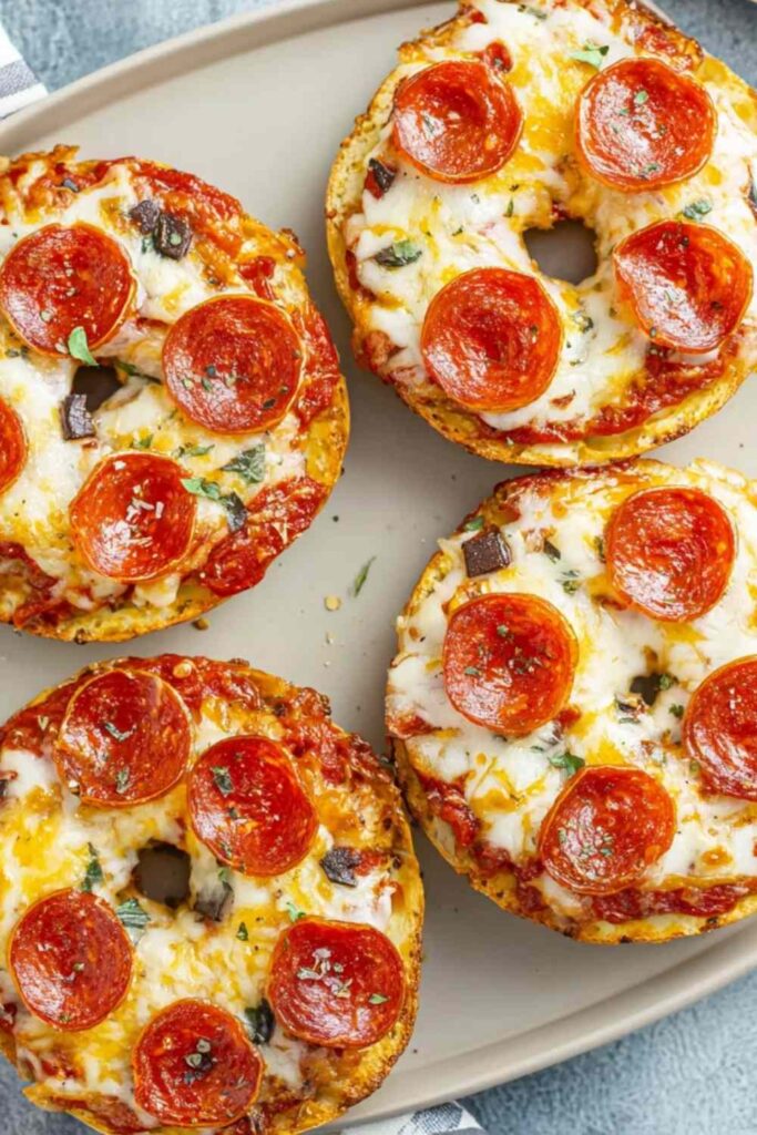 Baked mini pizza bagels on a metal tray, topped with melted cheese, pepperoni, and seasoning, with crisp golden edges.