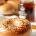 Golden baked New York-style bagel with sesame seeds in front of a jar of amber honey on a white cloth