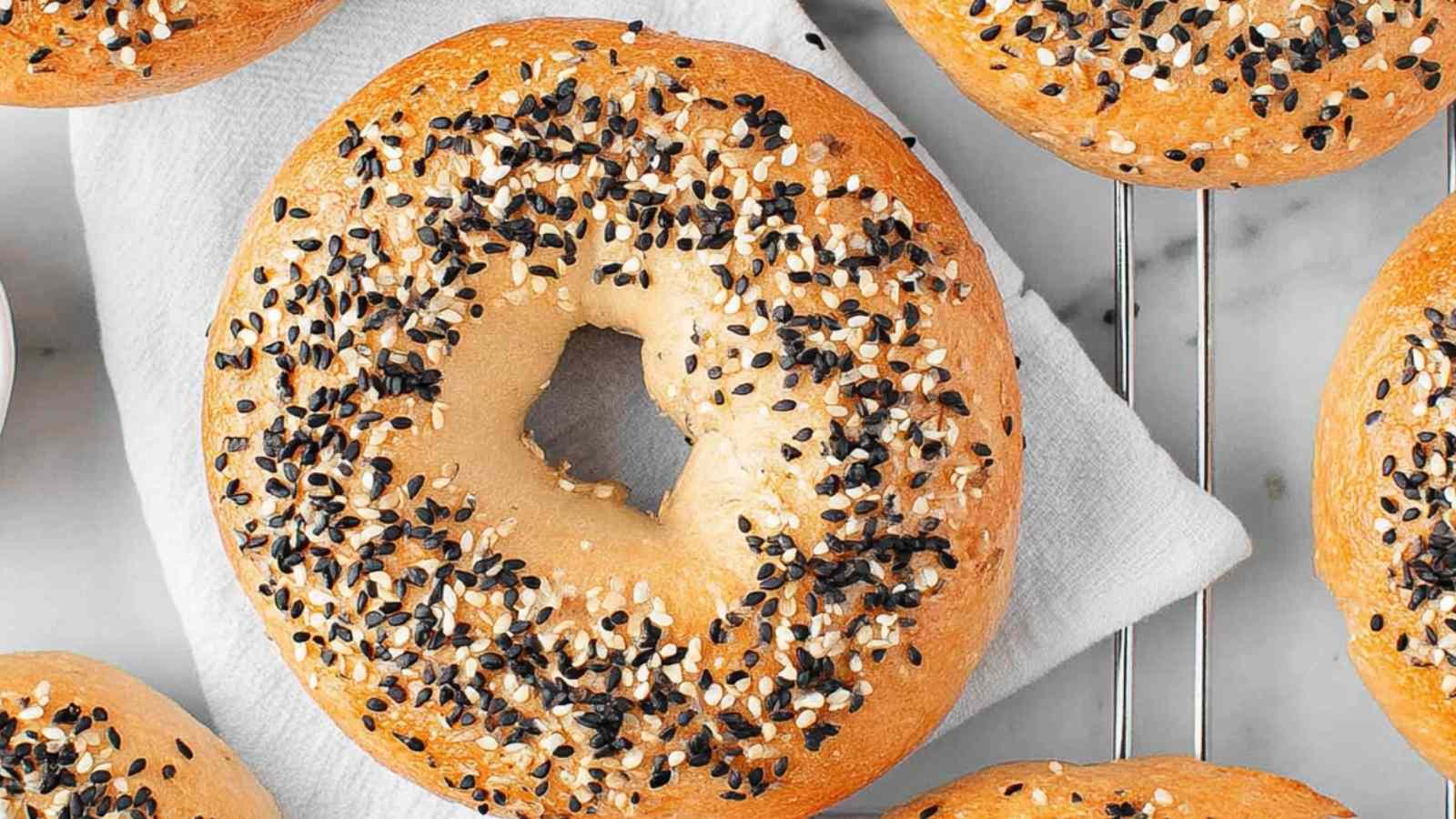 Close-up of a golden-brown sesame bagel topped with black and white sesame seeds resting on a cooling rack with a white towel underneath